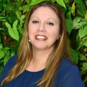 Smiling woman in blue dress with green leafy background.