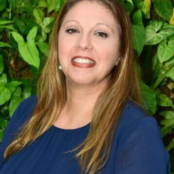 Smiling woman in blue dress with green leafy background.