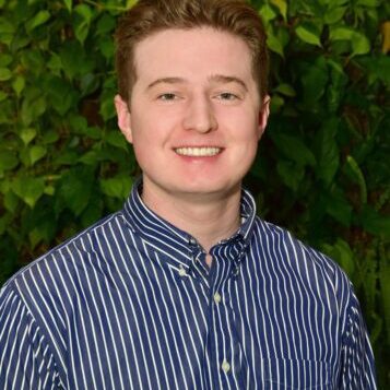 A young man smiling in front of leafy green background.