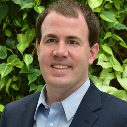 Man in a suit smiling outdoors with green foliage background.