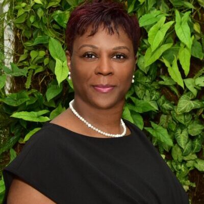 Professional woman in a black dress with a green leafy background.