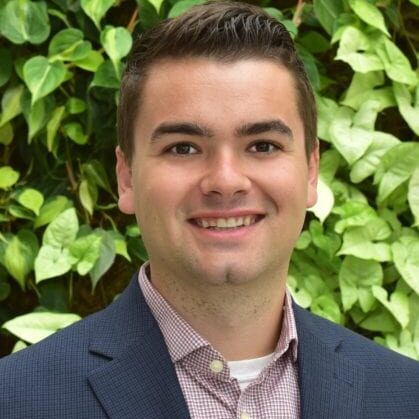 Young man in a suit smiling outdoors with green leafy background.