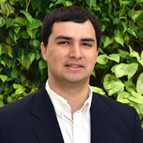 A man in a suit stands before a lush green background.