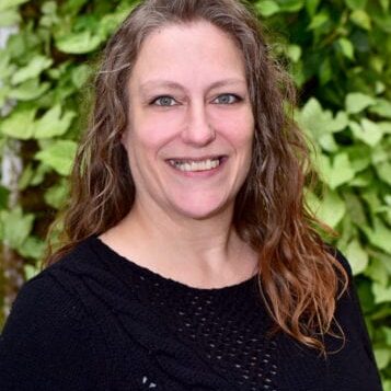 Smiling woman with wavy hair wearing a black top against green foliage.