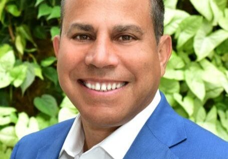 Smiling man in a blue suit with a leafy background.