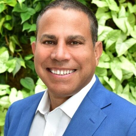 Smiling man in a blue suit with a leafy background.