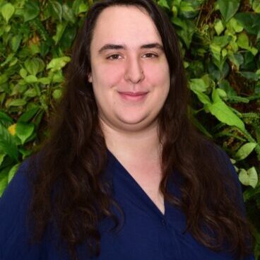 A person smiling with long dark hair against a leafy green background.