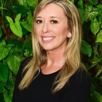 Smiling woman with blonde hair in front of green foliage.