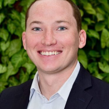 A smiling man in a suit with a leafy green background.