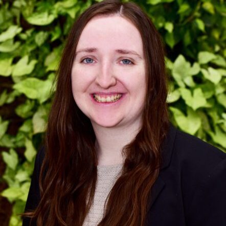 A woman with long brown hair smiling in front of green foliage.