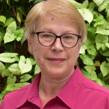 Portrait of a smiling woman with glasses and short blonde hair in front of green foliage.