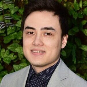 Young man in a gray blazer smiling against a leafy background.