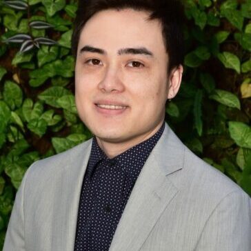 Young man in a gray blazer smiling against a leafy background.