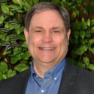 Smiling man in a blue shirt and dark blazer stands before green foliage.