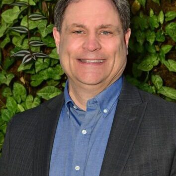 Smiling man in a blue shirt and dark blazer stands before green foliage.