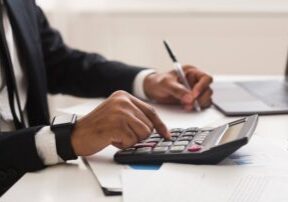 Person in a suit using a calculator and writing on papers at a desk with a laptop nearby.